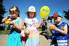 Ein Mädchen sprudeln durch einen Strohalm in ein Gefäß, in ihrer Mitte steht ein Mädchen mit Luftballon